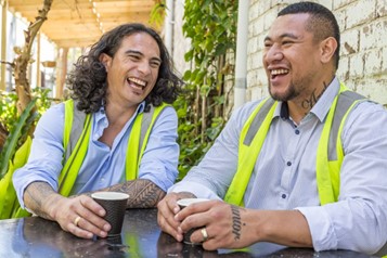 Two men in blue shirts and high vis vests sit at an outside table holding takeaway cups of coffee. They are both laughing.